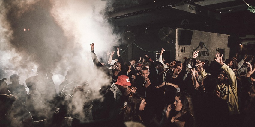 A crowd of people partying with smoke and raising their hands in the air while dancing.
