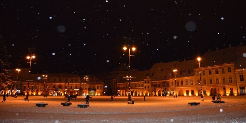 Sibiu’s grand square at night with snow falling, illuminated street lamps and people
