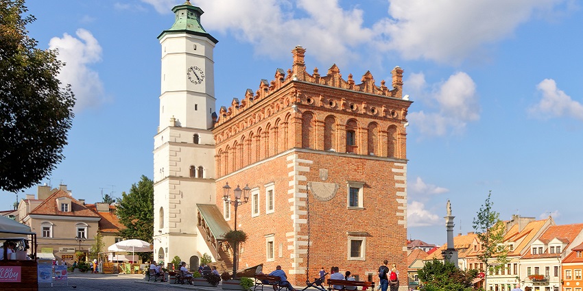 Sandomierz with a tall white clock tower, people sitting on the benches and walking around.