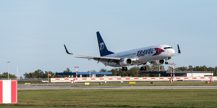 An aeroplane taking off from the runway with greenery and an airport complex visible