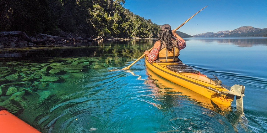 An image of a woman on a boat paddling on a calm river surrounded by serene views.