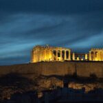 Acropolis Hill and Parthenon illuminated at night with ruined architecture and blue sky.