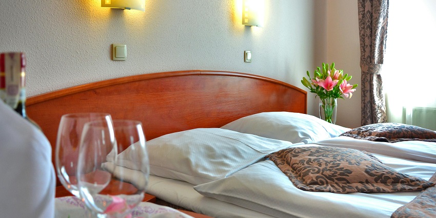 Clear wine glass on a wooden table beside a cosy bed in a softly lit room.