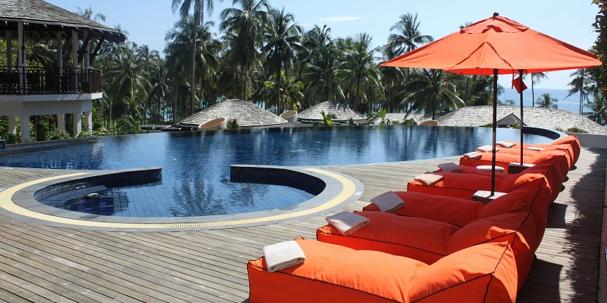 Poolside view of a lodge with seating area, an amazing ambience, and many coconut trees around it.