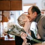 A couple kissing while the woman holding a flower bouquet with a wine bottle and a glass