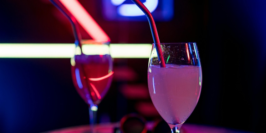 Two cocktails glasses coloured pink served with straws and neon lights in the background