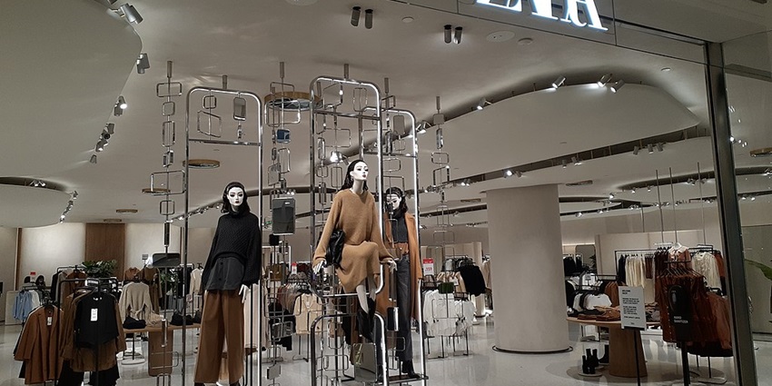 Zara store with dressed mannequins and clothes on display with a lit Zara sign outside.