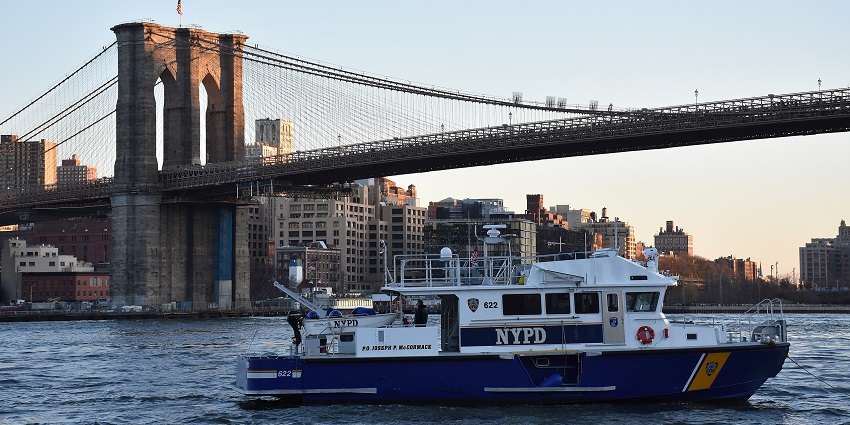 An image of an NYPD police boat on the water with the Brooklyn Bridge and Downtown Brooklyn at sunset.