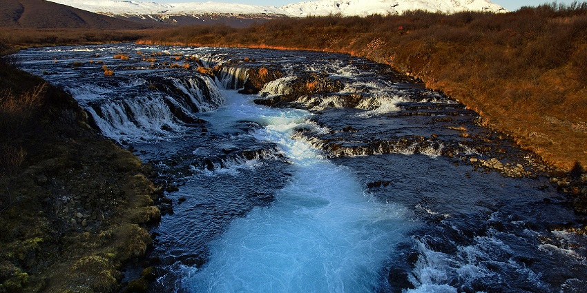 An image of Bruarfoss waterfall with clear blue water flowing over rocks and surrounded by greenery.
