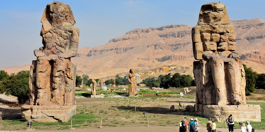 Colossi of Memnon with tall old ruined statues, people visiting and mountains behind