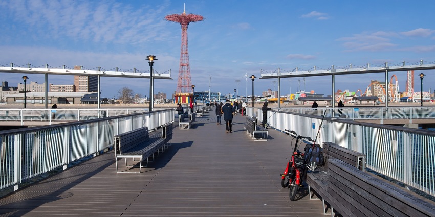 Coney Island boardwalk with visitors, ocean view, and colorful amusement rides.