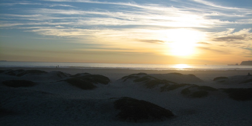 Rocks are partially covered in sand at Coronado Beach, with two people enjoying the sunset