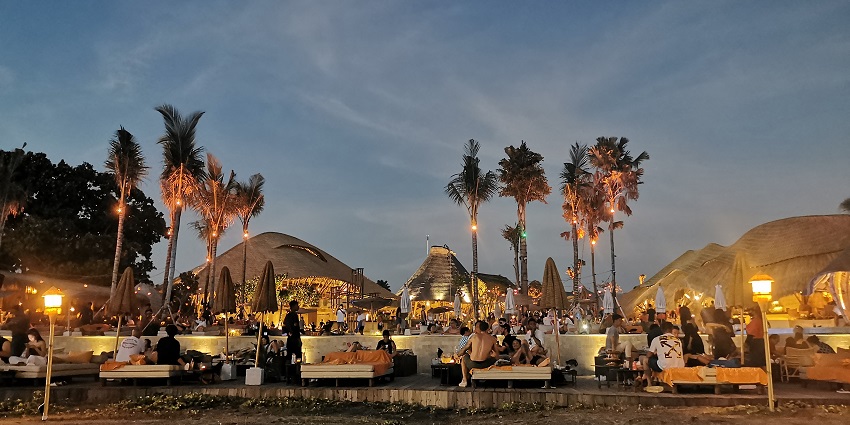 Beach club at night with lights on palm trees and people sitting on the lounge chairs.