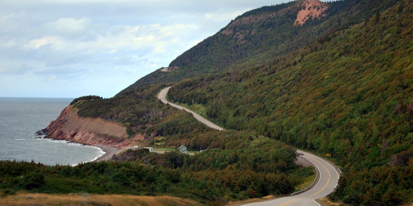 Cabot trail is surrounded by greenery with a sea on the one side and hills on the other side.