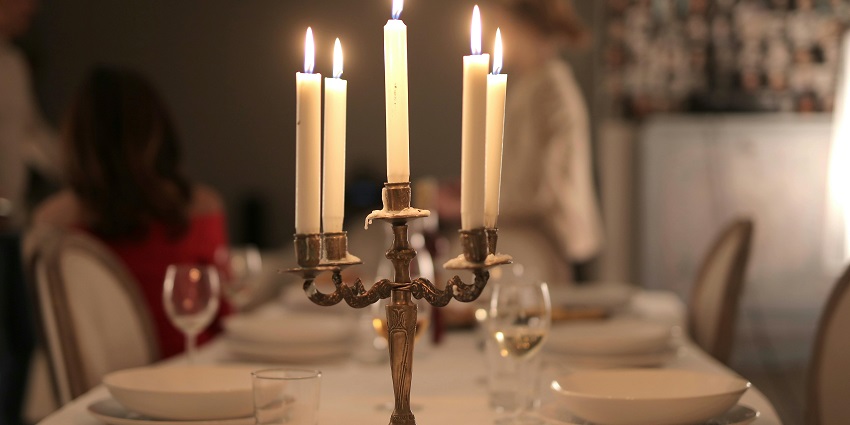 Two people's hands were placed on the dinner table with candles lit next to bread and glasses.