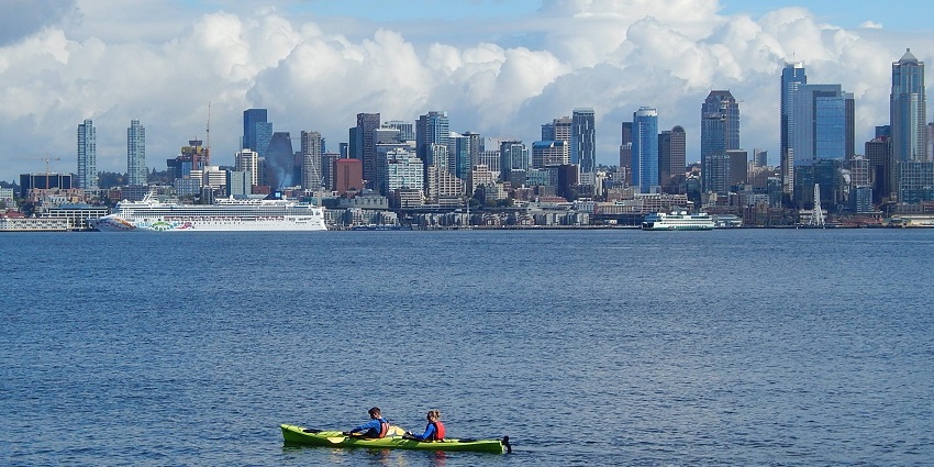 Two people kayaking across the calm waters of the bay and tall buildings in the background