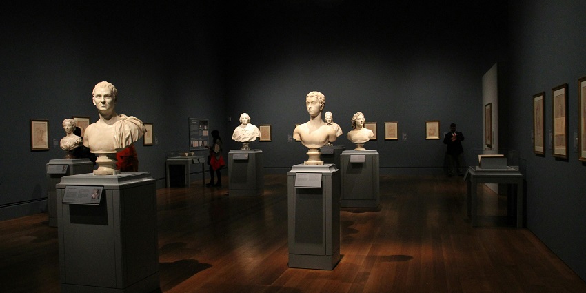 Assorted range of busts of popular people lined up on a stand in a dark room of a museum.