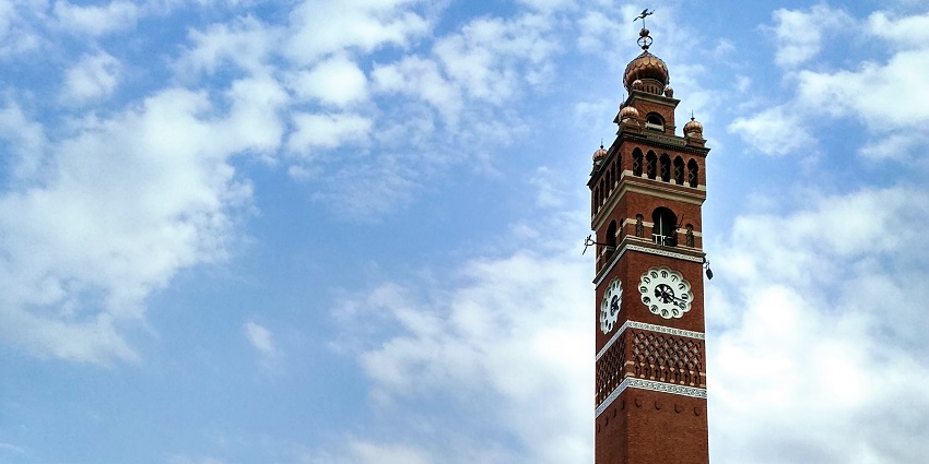 An image of Clock Tower in the centre of the road with a few cars and men with bicycles around.