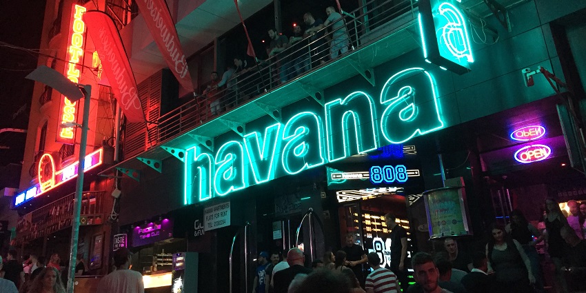 A busy street with the entrance to the Havana Malta club and people walking around
