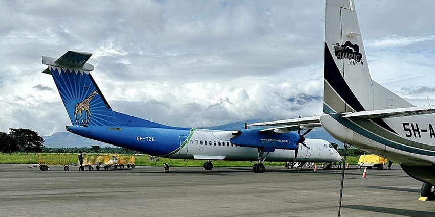 Aeroplanes parked at Arusha airport with a yellow van, trolleys and people standing.