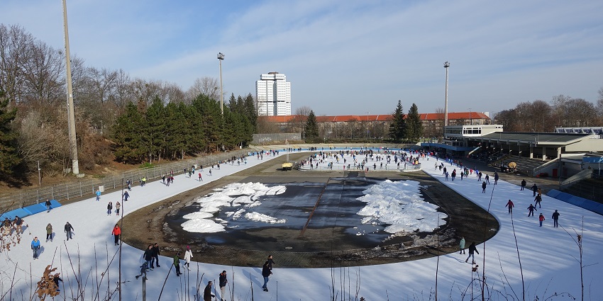 People ice skating in the Horst Dohm Eisstadion rink with big light poles and trees