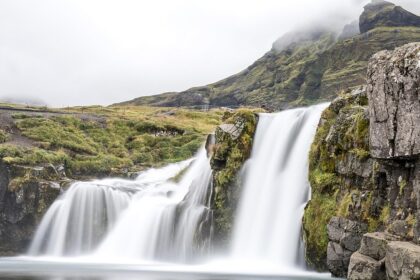 An image of a beautiful Iceland waterfall flowing over rocky hills, under the cloudy sky.