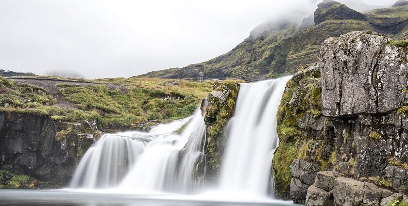 An image of a beautiful Iceland waterfall flowing over rocky hills, under the cloudy sky.