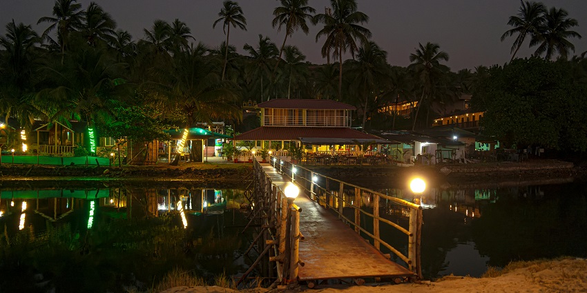 Vibrantly lit tropical resort buildings at night with a wooden bridge over the water.