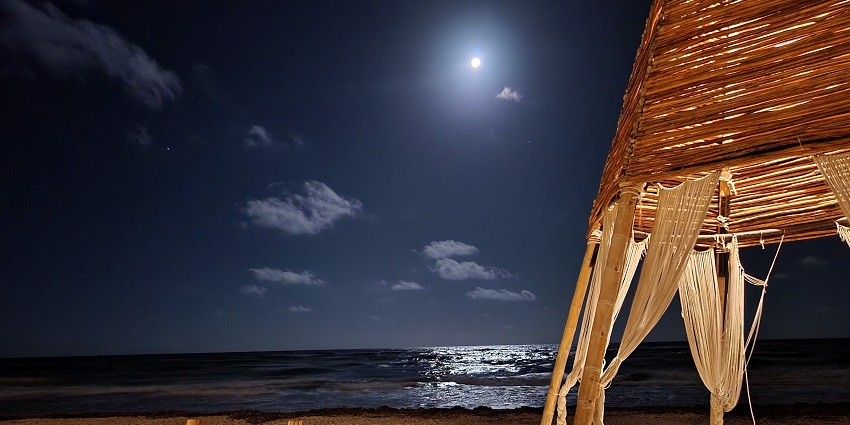 Beach club with lounge chairs and a tent with dark sea glowing below the bright moon