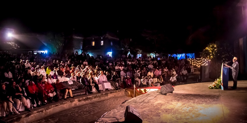 Speaker on stage addressing a large audience sitting on the stairs and pavement