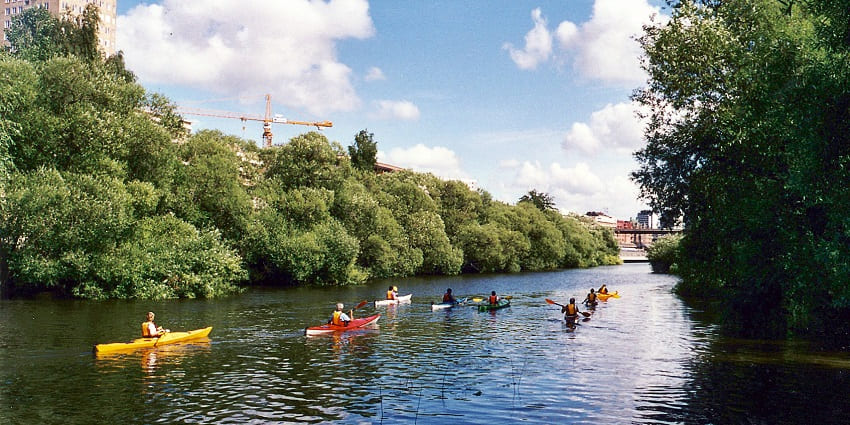 People kayaking solo on a Stockholm river with greenery reflecting on the calm waters