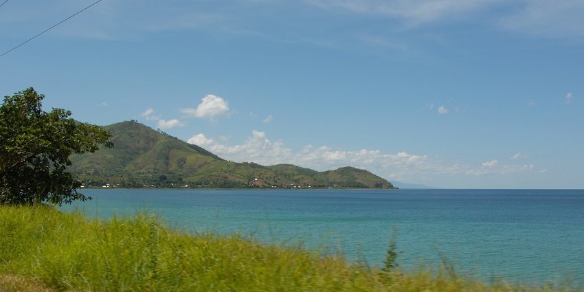 Lake Tanganyika Beach with a sandy shoreline and people walking under a blue sky and trees