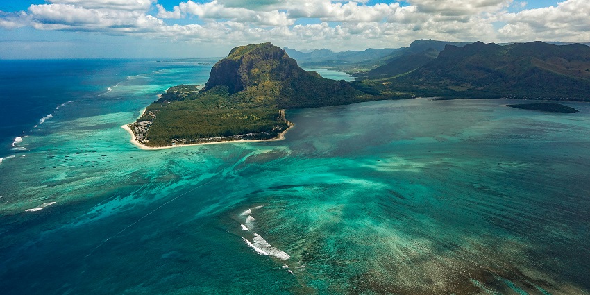 Le Morne peninsula beach with the sandy shoreline, tall palm trees and distant buildings.