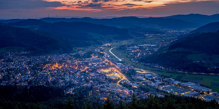 City buildings along a road with hills in the distance, lit by a sunset sky.