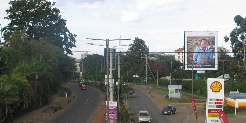 Langta Road with vehicles passing by, a petrol pump on one side, a poster and trees.