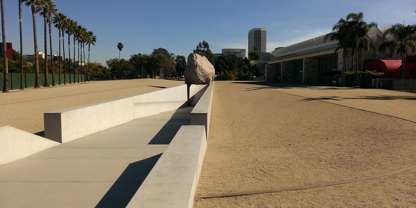 A large boulder suspended above a walkway at LACMA, with visitors walking underneath.