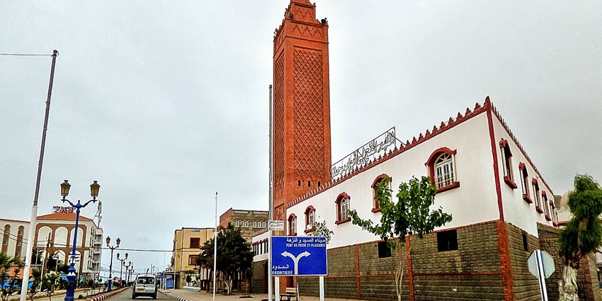 Marsa mosque in Malta with a tall red minaret, a car on the road and a signboard for directions.