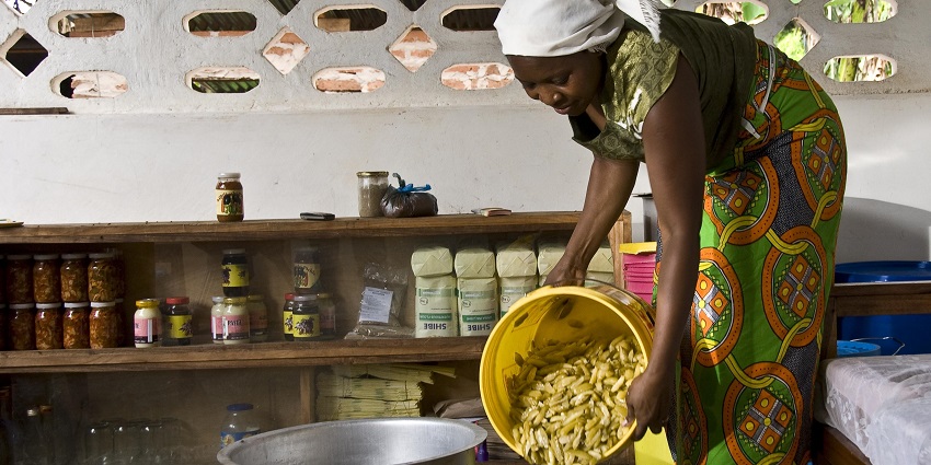 A woman pouring and preparing pickles in big containers and small pickle jars on shelves .