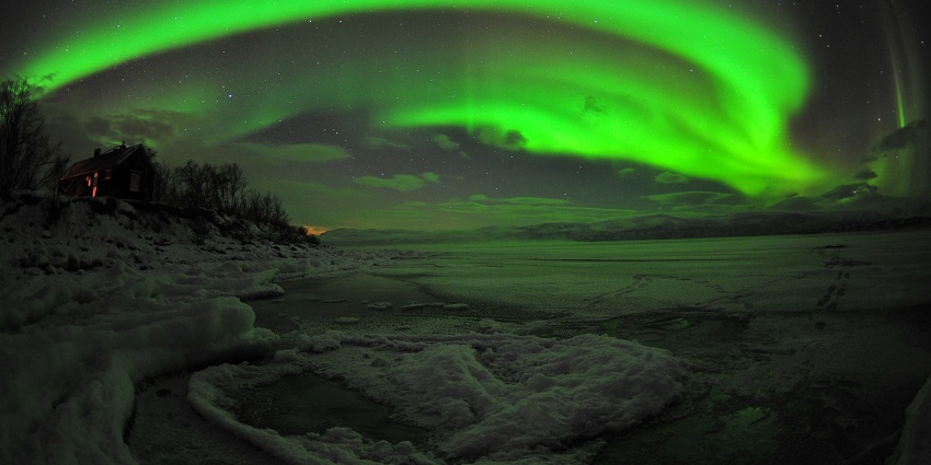 Northern lights are visible from Abisko with a small cabin surrounded by trees on the left