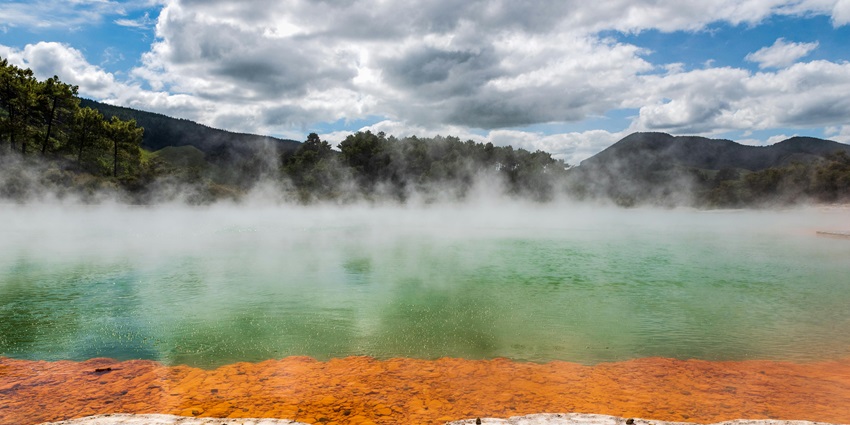 Hot spring with warm heat coming from it and surrounded by greenery under a cloudy sky.