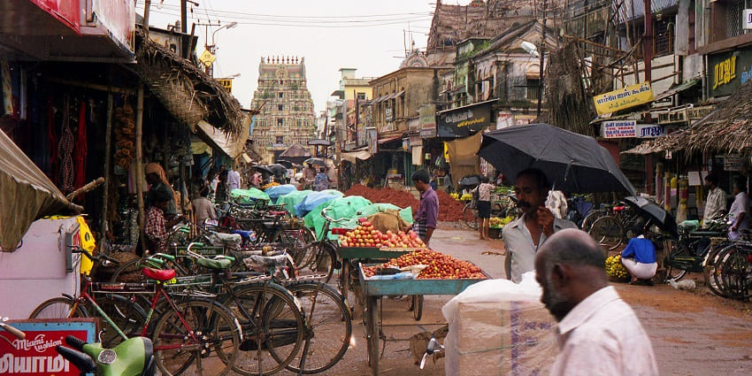A busy street in Kumbakonam with street vendors and bicycles parked in rainy weather.