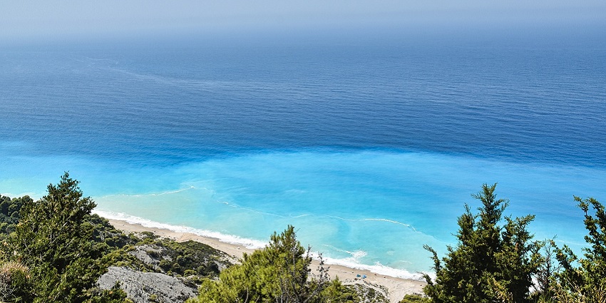Scenic beach with turquoise water, rocky cliffs, and greenery under a clear blue sky.
