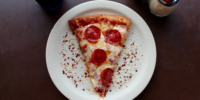 Image of a Pizza slice served on a white plate with oregano and chilli flakes on the side