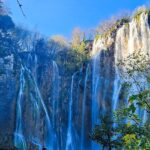 Text: A view of extremely beautiful Plitvice Waterfalls cascading behind the lush green trees.