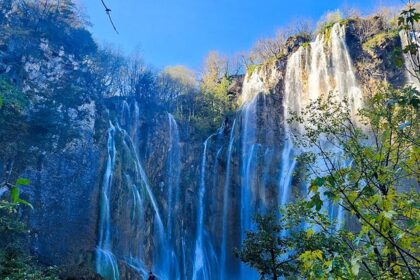 Text: A view of extremely beautiful Plitvice Waterfalls cascading behind the lush green trees.