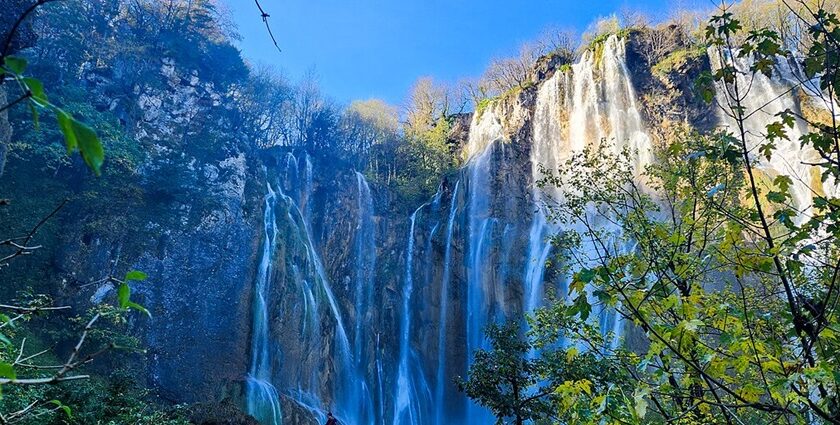 Text: A view of extremely beautiful Plitvice Waterfalls cascading behind the lush green trees.