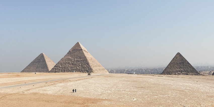 Two men are walking towards the pyramids of Giza on a sunny and dry morning through sands.