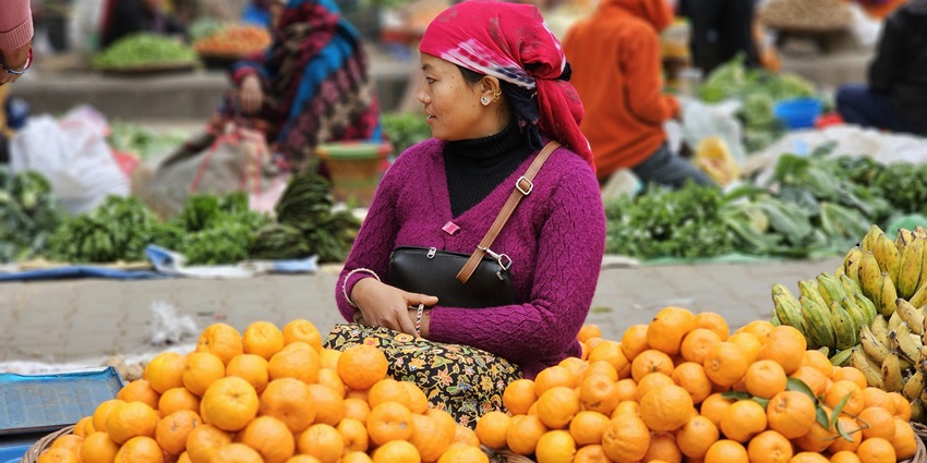 A woman selling root crops wearing a pink sweater and a man standing in the background.