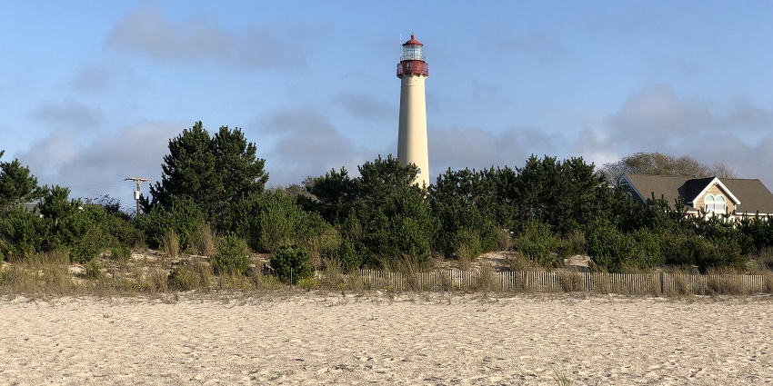 Cape May beach with sand, a fence, green trees and Cape May lighthouse in the background