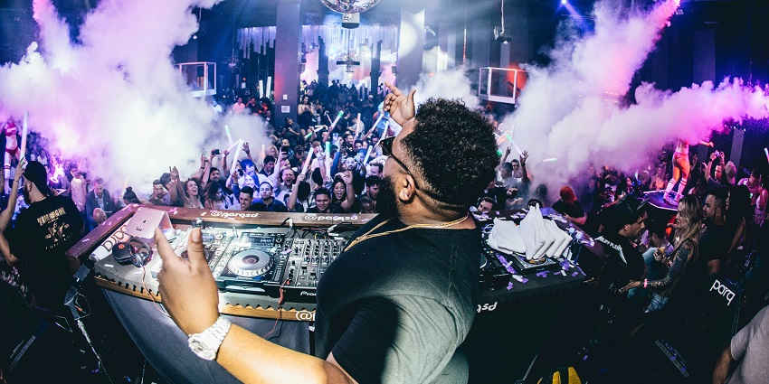 A scene in a club with DJ playing music, people dancing and visual effects lighting.